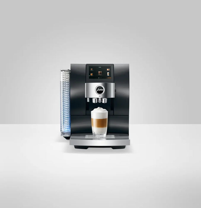JURA Z10 Aluminium Dark Inox Signature Line EA volautomatische espressomachine met verlicht waterreservoir, touchdisplay en vers bereide latte macchiato in een glas op de lekbak, frontaal weergegeven tegen een grijze achtergrond